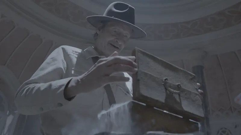 Filmstandbild ungegradet: Mann mit Hut beugt sich über eine Kiste, flache, graue Farben