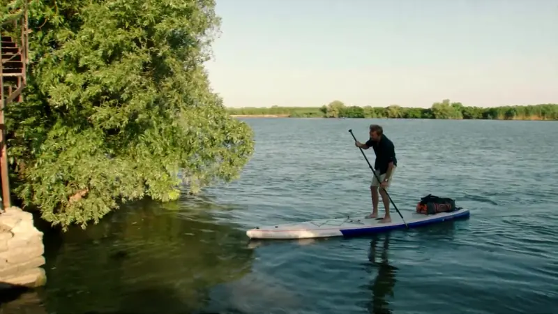 Stand-up-Paddler auf der Donau neben einem Baum am Ufer – Dokumentarfilm Ein Mann. Ein Fluss. Ein Abenteuer